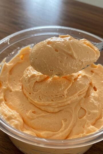 In our home, fluff desserts were always one of those clever “sneaky” treats my mom would make, packing in a little extra nutrition without anyone being the wiser. Her recipes always included cottage cheese, which meant our sweet indulgences were secretly full of protein. She would whip up these fluff-style desserts to keep in the fridge for quick weeknight treats. With their simplicity and minimal ingredients, they were the perfect solution for a busy mom juggling work and family. Although she made a variety of flavors—from lemon to strawberry-banana to pineapple—orange fluff has always been my personal favorite. White bowl filled with orange fluff set on a white tray. Cottage Cheese…In a Dessert? If you already love cottage cheese, you’re sold. If not, let me convince you. This orange fluff does not taste like cottage cheese—I promise! The secret is to puree the cottage cheese until it’s completely smooth. It adds a subtle tang and creamy texture to the dessert, but all you’ll notice is the luscious orange flavor. Three small white bowls filled with orange fluff, garnished with whipped cream and fresh orange slices. There’s something so delightful about a dessert that mimics my favorite ice cream—creamsicle—without much effort. Total win, if you ask me. (It’s also why I adore orange fruit dip!) When I went away to college, I worked as a server and actually saved up my tip money for a week to buy an inexpensive food processor from Meijer—just so I could have fluff whenever I wanted. That’s serious dedication, and it shows just how much I adore my mom’s sneaky orange fluff. Ingredients for orange fluff arranged on a beige countertop. How to Make My Mom’s Orange Fluff Orange fluff is one of the simplest desserts to prepare. That’s why Mom made it so often, but it’s also delicious enough to add to any holiday dessert table. Ingredients You’ll Need This recipe uses four basic ingredients plus one optional addition: Cottage cheese: Adds protein, a slight tang, and creamy texture. Orange Jell-O mix: Infuses the fluff with sweetness and orange flavor. Thawed whipped topping: Creates the airy, fluffy texture. Use Cool Whip, TruWhip, or stabilized whipped cream. Canned mandarin oranges: Provides sweet, juicy orange pieces in every bite. Optional – miniature marshmallows: Adds extra texture and sweetness if desired. Making the Recipe Puree the cottage cheese: Use a food processor to blend until completely smooth. Mix in Jell-O: Transfer the pureed cottage cheese to a bowl and whisk in the dry orange Jell-O mix until fully incorporated. Fold in whipped topping: Use a rubber spatula to gently fold in the thawed whipped topping. Add fruit and extras: Fold in mandarin oranges and marshmallows, if using. Chill: Cover the orange fluff and refrigerate for at least 2 hours to allow flavors to meld and the texture to set. Serve: Garnish with whipped cream and fresh orange slices for a pretty presentation. Chopped mandarin oranges being added to a bowl of orange fluff. FAQs Can I use fresh oranges instead of canned mandarin oranges? Canned mandarins are convenient since they’re peeled and ready to use. Fresh oranges can be used, but you’ll need to supreme them to remove pith and membrane. Fresh oranges are slightly less sweet than mandarins in syrup, so the final fluff may taste a bit less sweet. Can I make orange fluff ahead of time? Yes! The fluff needs at least 2 hours in the fridge, so you can prepare it a day or two in advance. It will keep for up to 3 days. Can I freeze this orange fluff? Freezing is not recommended. Cottage cheese can separate when frozen and thawed, which may leave the fluff watery and less creamy. Can I use other Jell-O flavors? Absolutely! One of the best things about Mom’s fluff recipes is their versatility. Swap flavors of Jell-O and fruit to create your own variations—for example, pineapple fluff uses pineapple Jell-O and canned pineapple. Orange Fluff A light, dreamy dessert that comes together in minutes and is perfect for any occasion! Creamy cottage cheese blends with orange Jell-O and whipped topping for a sweet, citrusy treat. Prep Time: 10 minutes Chill Time: 2 hours Total Time: 2 hours 10 minutes Servings: 10 Equipment: Food processor Large mixing bowl Rubber spatula Ingredients 16 oz cottage cheese 2 boxes orange Jell-O mix (3 oz each) 16 oz frozen whipped topping, thawed 2 cans mandarin oranges (11 oz each), drained and roughly chopped 2 cups miniature marshmallows (optional) Instructions Puree cottage cheese in a food processor until smooth. Transfer to a large bowl and whisk in dry Jell-O mix until fully incorporated. Fold in whipped topping with a rubber spatula until no streaks remain. Gently fold in mandarin oranges (and marshmallows, if using). Cover and refrigerate for at least 2 hours. Serve chilled, optionally topped with extra whipped topping and fresh orange slices. Notes Adding miniature marshmallows gives extra texture and sweetness. Orange Fluff